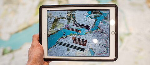 Eine Hand hält ein Tablet über eine Landkarte und zeigt mithilfe von Augmented Reality weitere Inhalte.